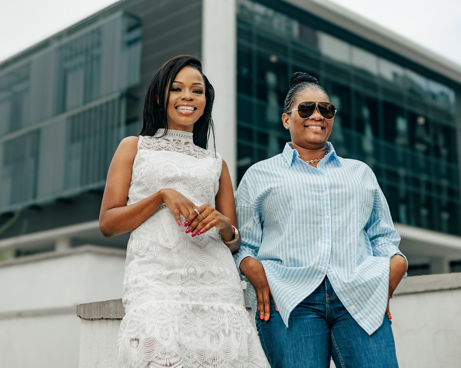 Two women in casual and elegant attire smiling in front of a modern building.