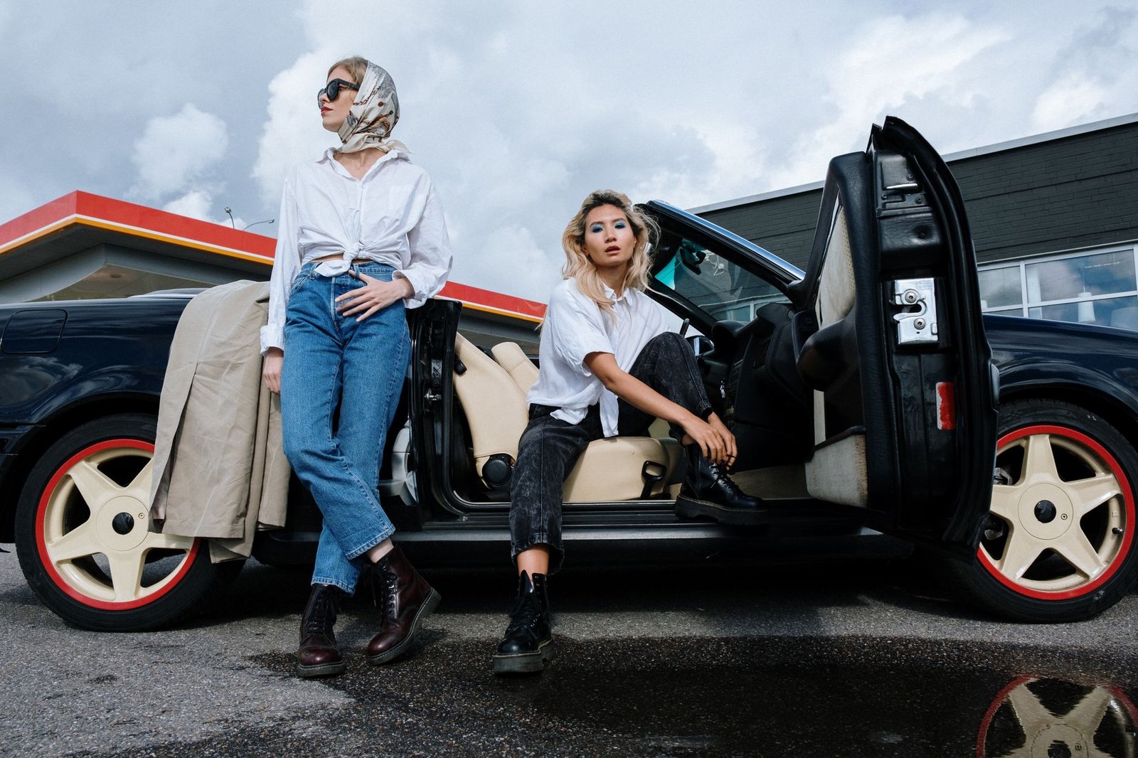 Two fashionable women posing with a convertible car outside a gas station.