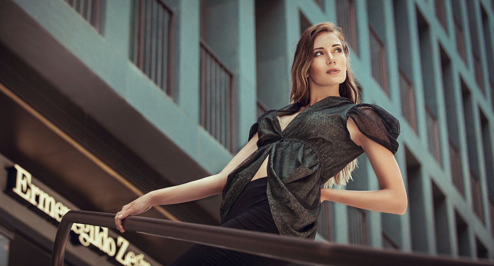 Fashion model in black blouse posing by urban railings, city setting.