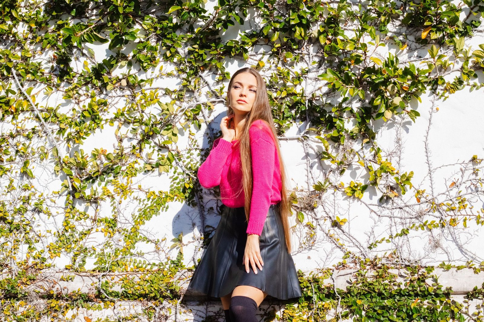 Woman in pink sweater and leather skirt poses against ivy-covered wall, showcasing modern style.