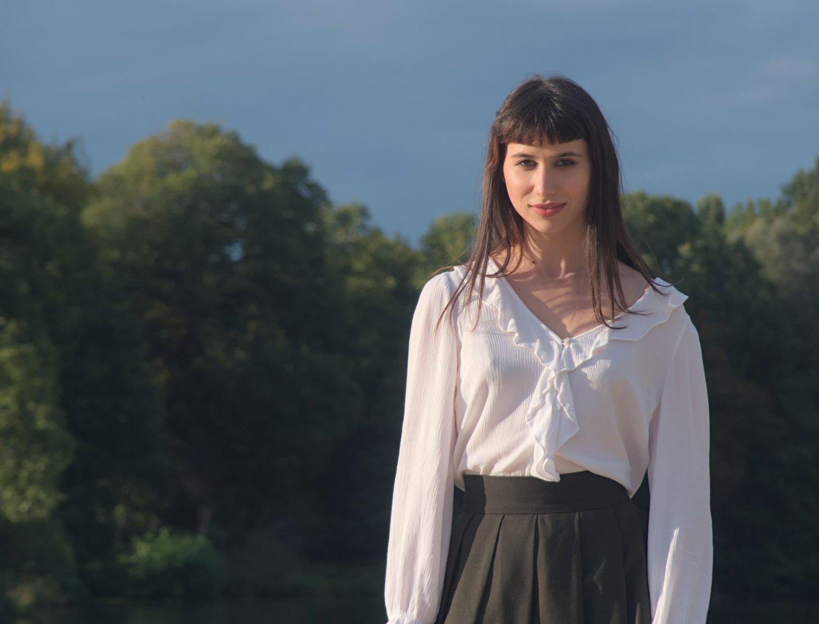 Young woman in a ruffled white blouse and skirt posing outdoors against a lush park backdrop.