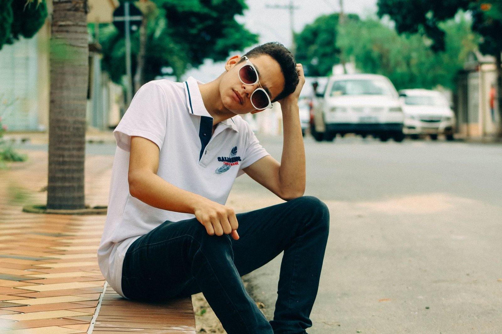 Casual young man sitting on a sidewalk by the road wearing sunglasses and a polo shirt.