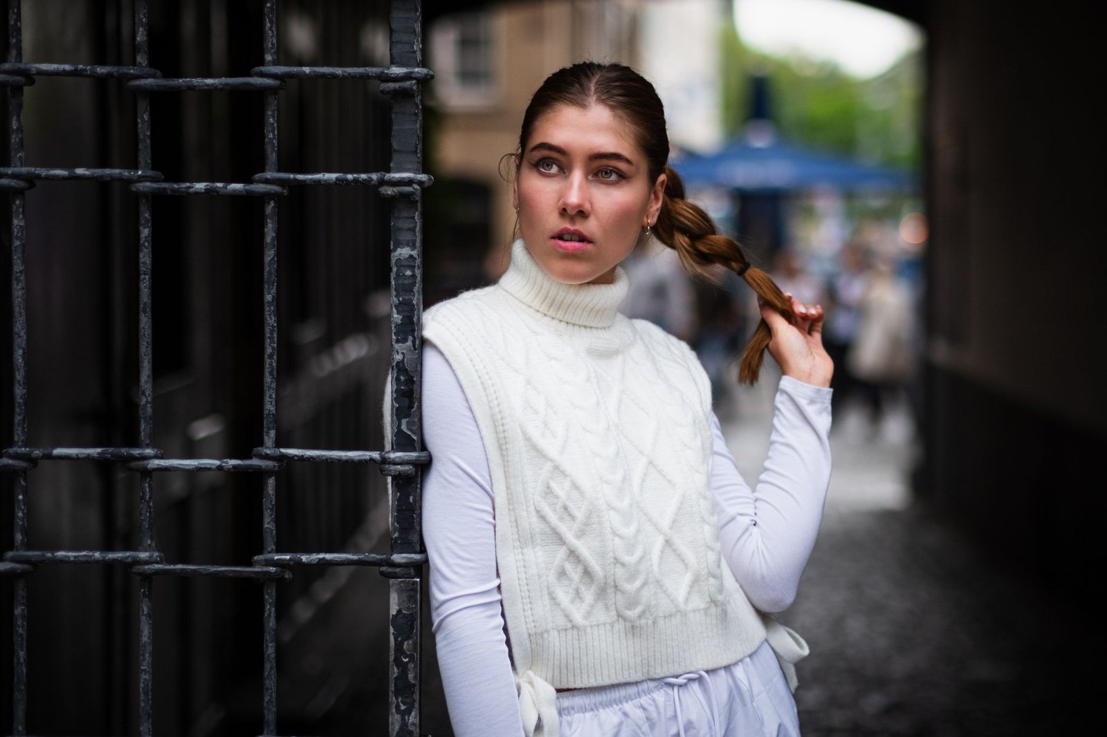 Woman in white turtleneck vest with braided hair standing by gate outdoors.