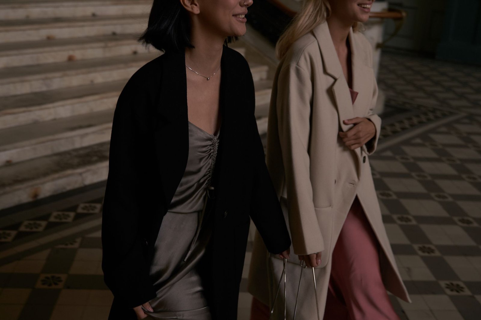 Two women wearing stylish coats walk indoors on patterned tiled fl or, exuding elegance.