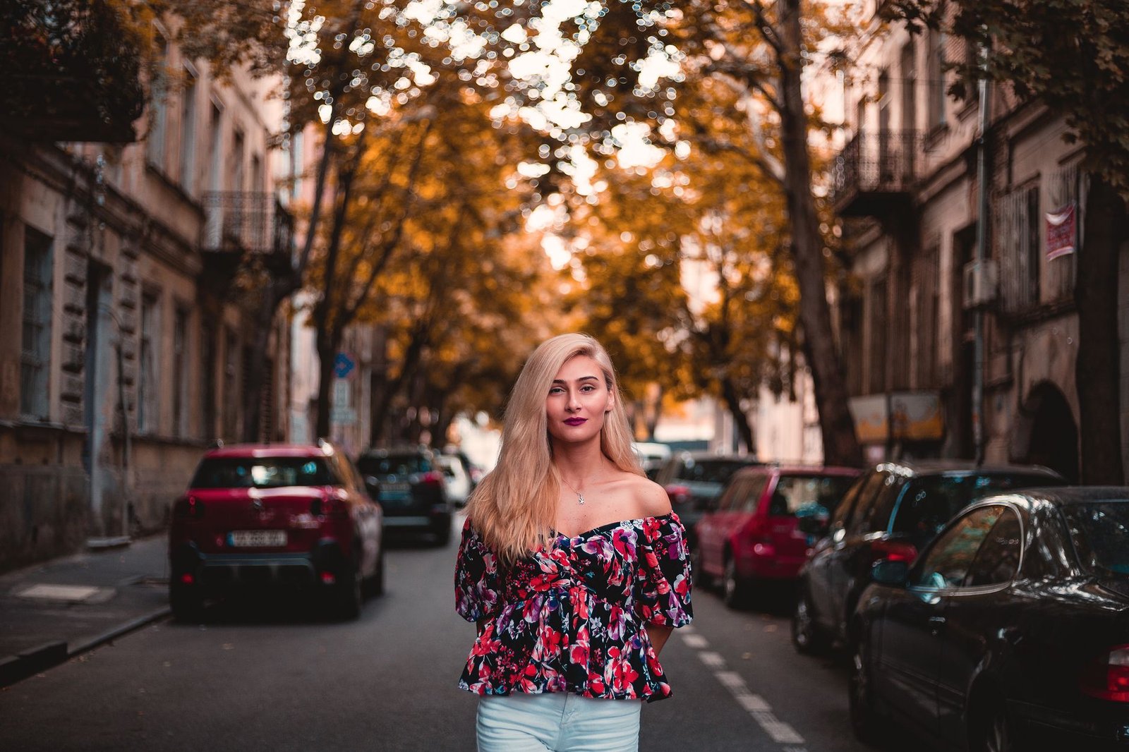 Stylish blonde woman wearing a floral top standing on a tree-lined urban street.