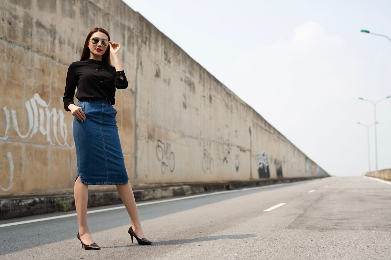 Fashionable woman in denim skirt and sunglasses posing confidently on urban street.