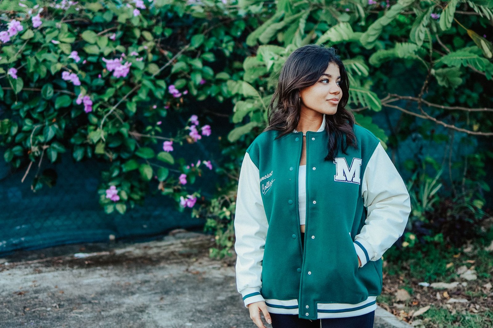 Woman in a green varsity jacket standing in a lush garden with lilac flowers.