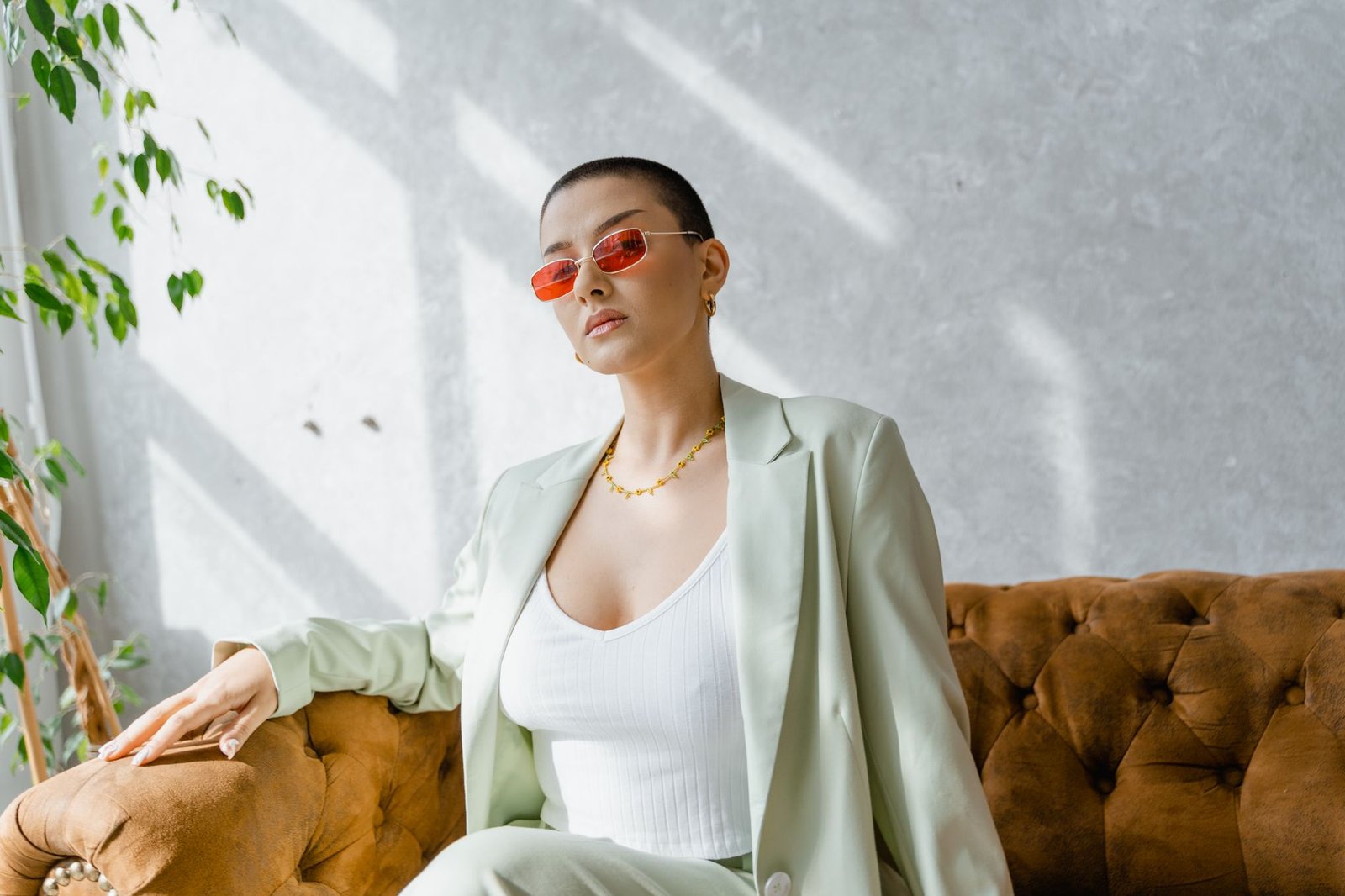 Confident woman in red sunglasses and chic suit sitting on a vintage couch in a sunlit room.