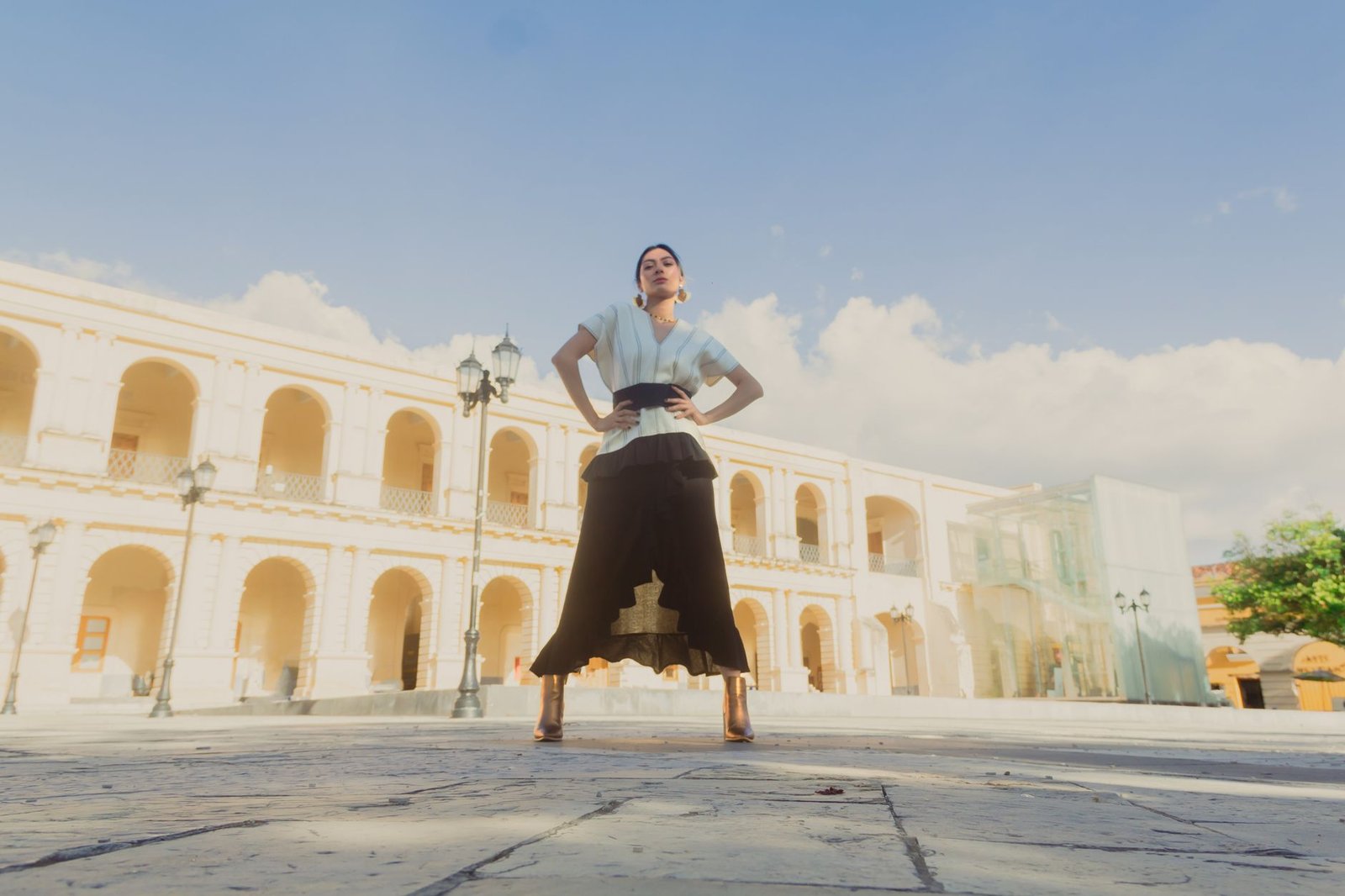 Elegant woman poses stylishly in a historical courtyard with clear blue sky.