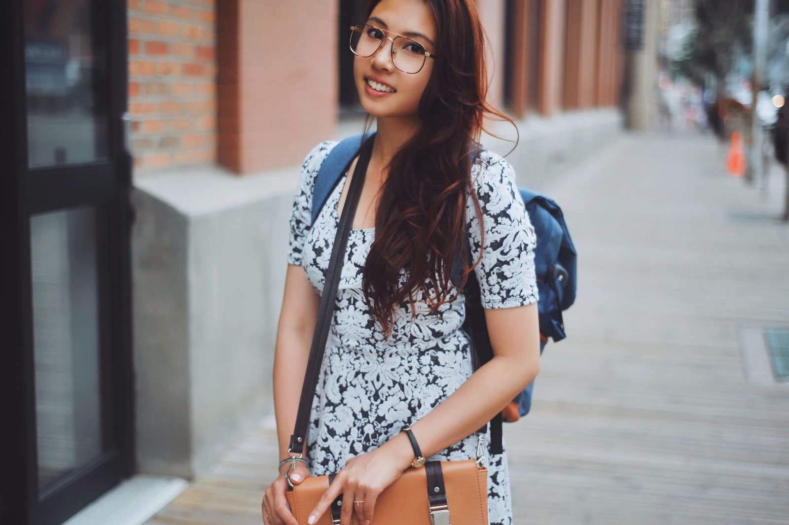 Trendy young woman in a floral dress and glasses smiling on a city sidewalk.