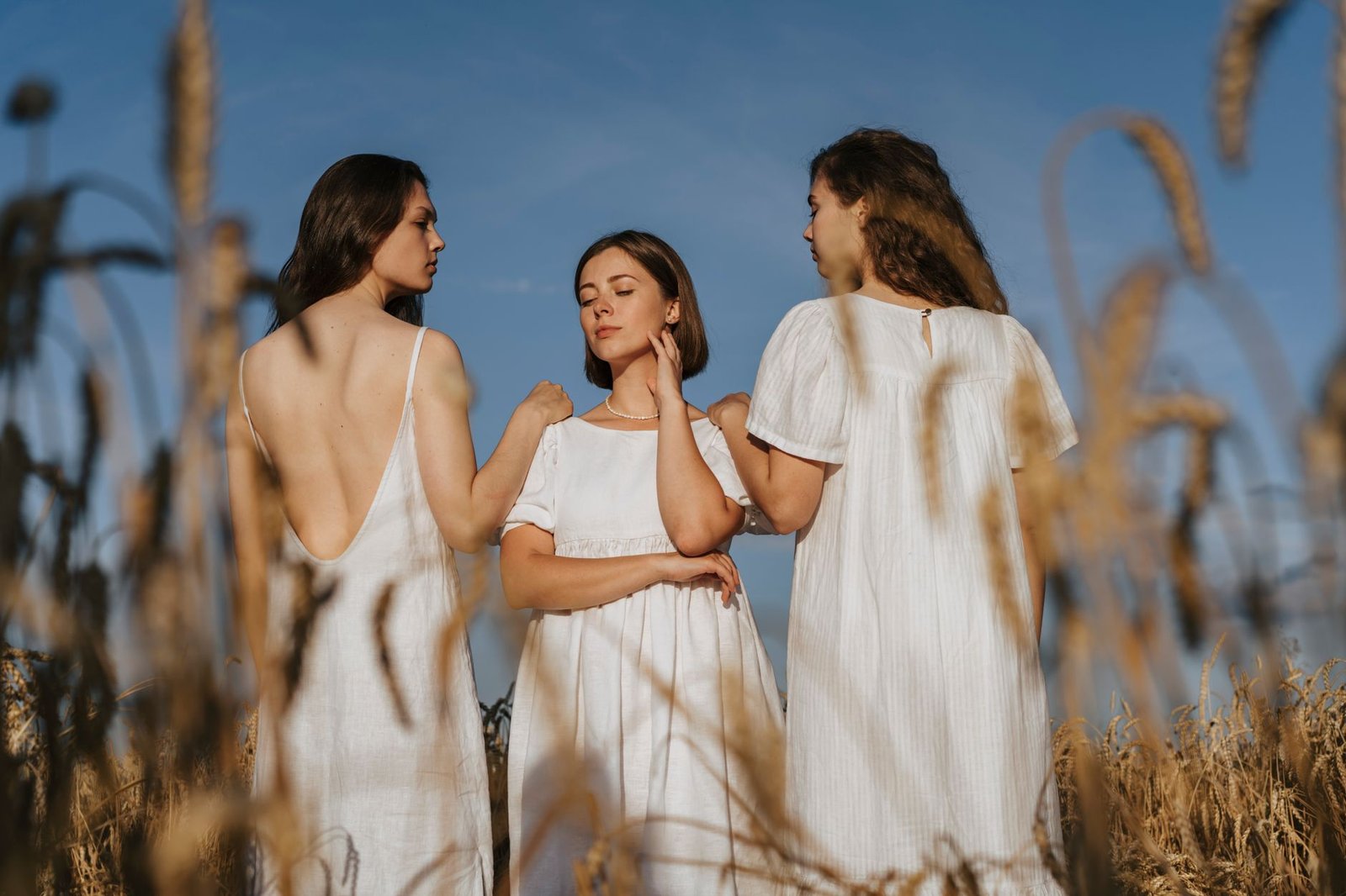 Three women in white dresses stand elegantly in a wheat field under a blue sky.