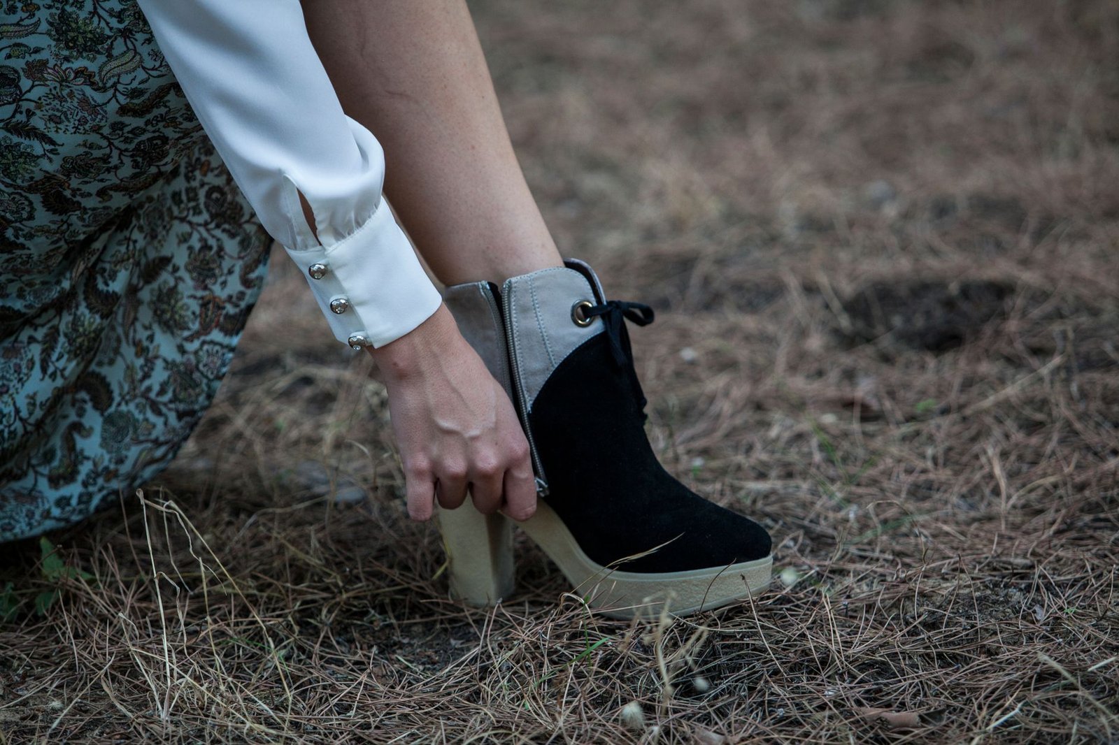 A woman adjusts her fashionable black boots while seated on the ground outdoors in fall.