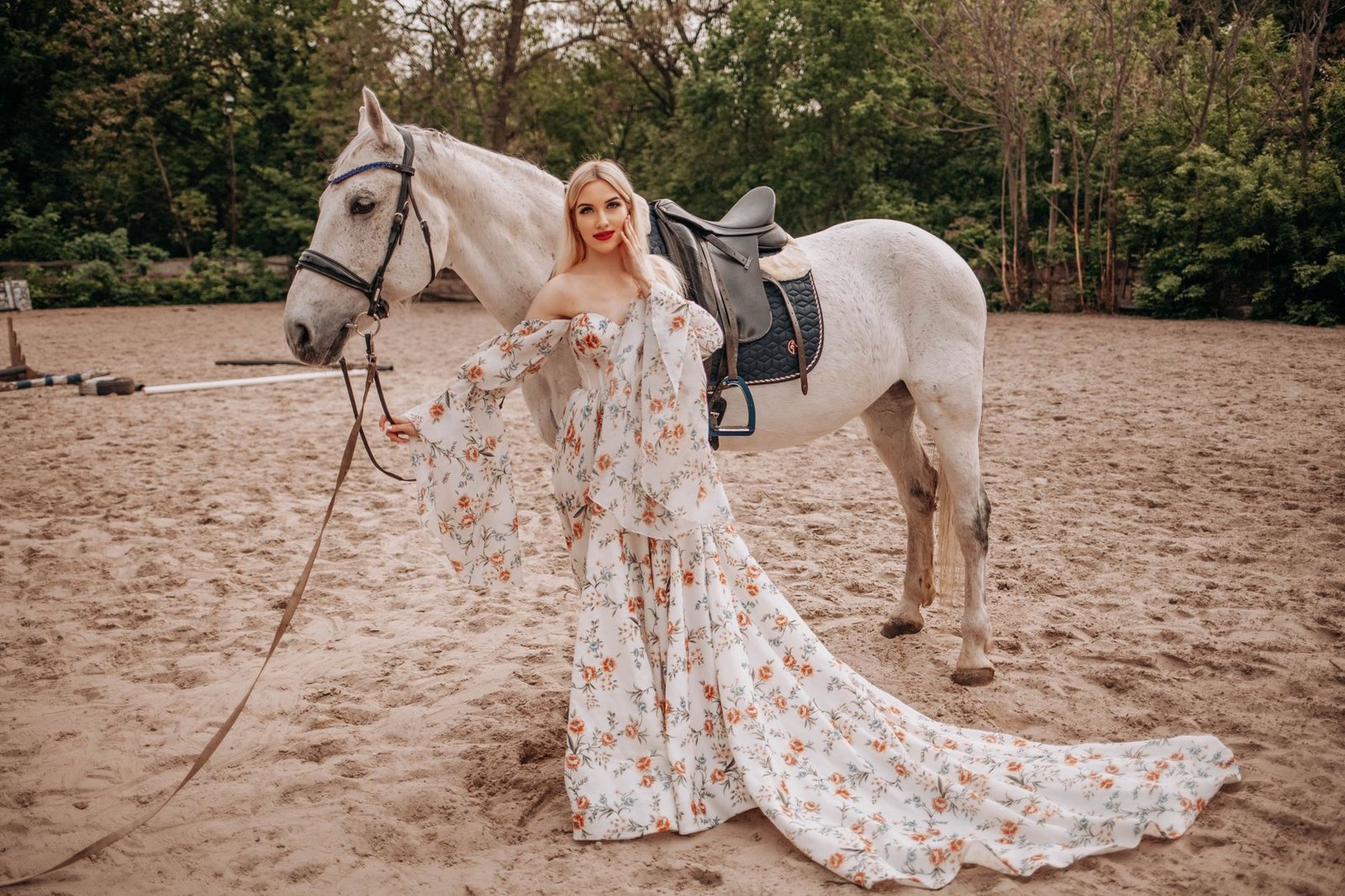 A stylish woman poses with a horse in an elegant floral dress on a sandy path.