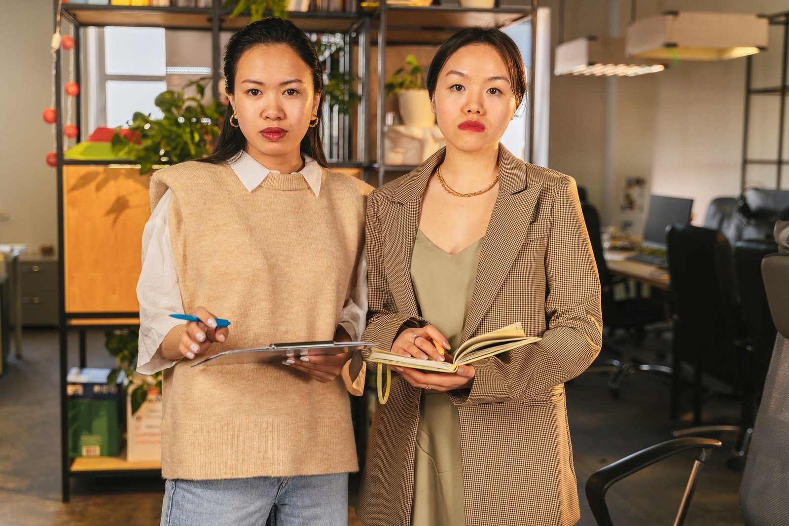 Two professional women standing in a modern office environment, holding documents and notebooks.