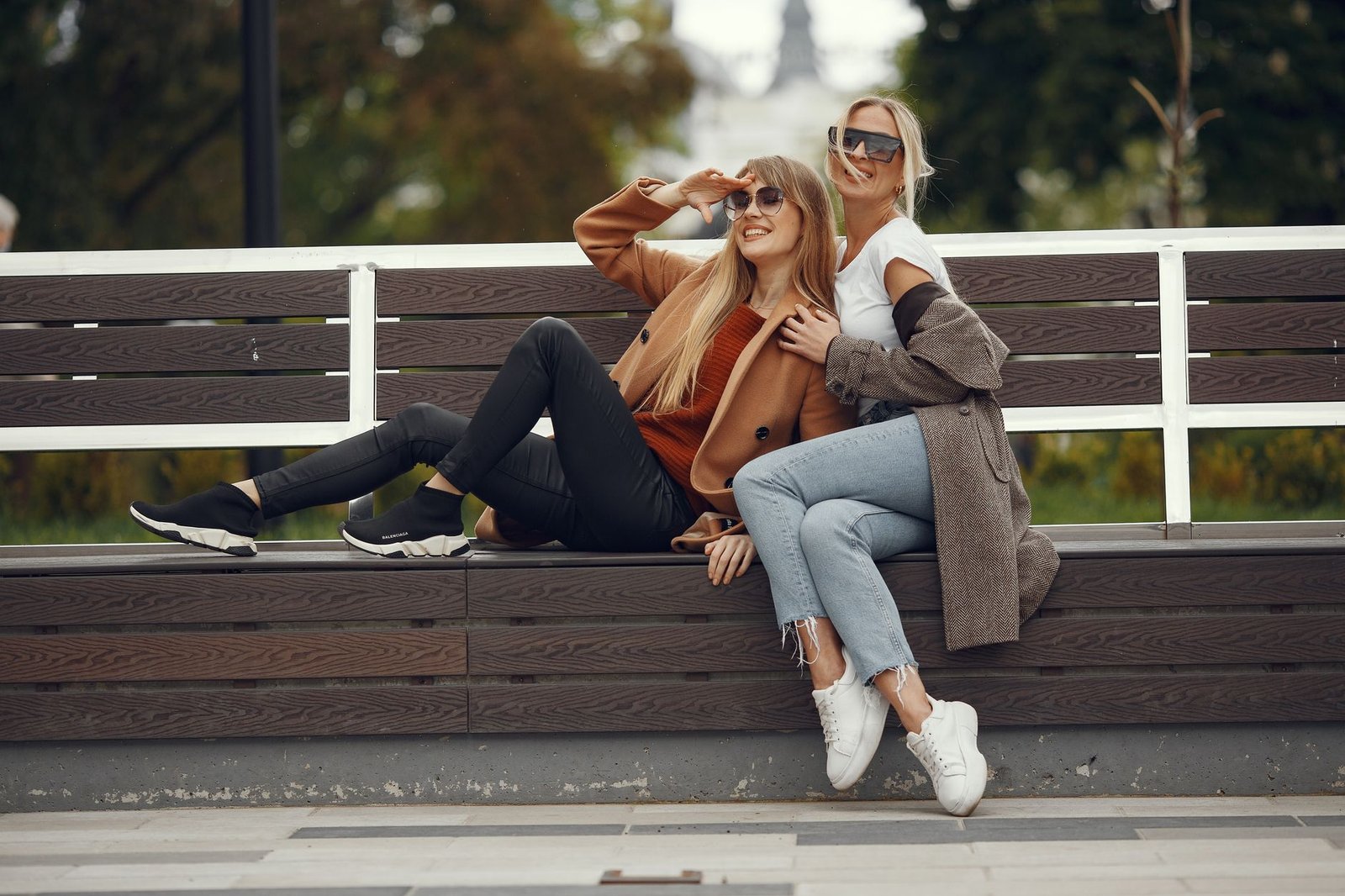 Two stylish women sitting and smiling on a bench outdoors in an autumn setting.