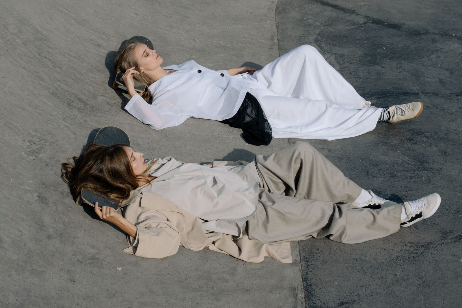 Two stylish women lying on skateboards at an outdoor skate park, dressed in trendy outfits.