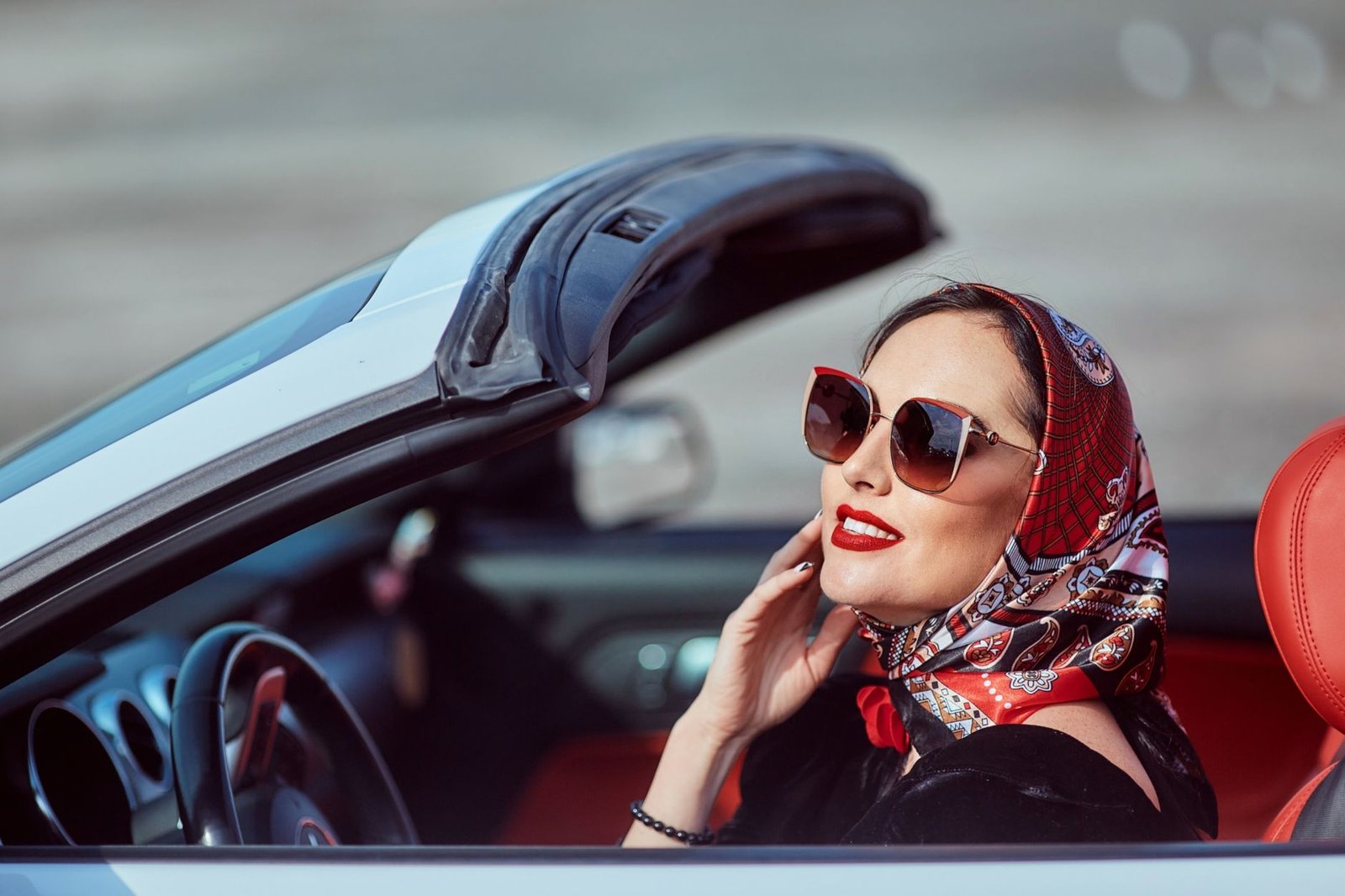 Stylish woman with sunglasses and headscarf enjoys a drive in a convertible on a sunny day.