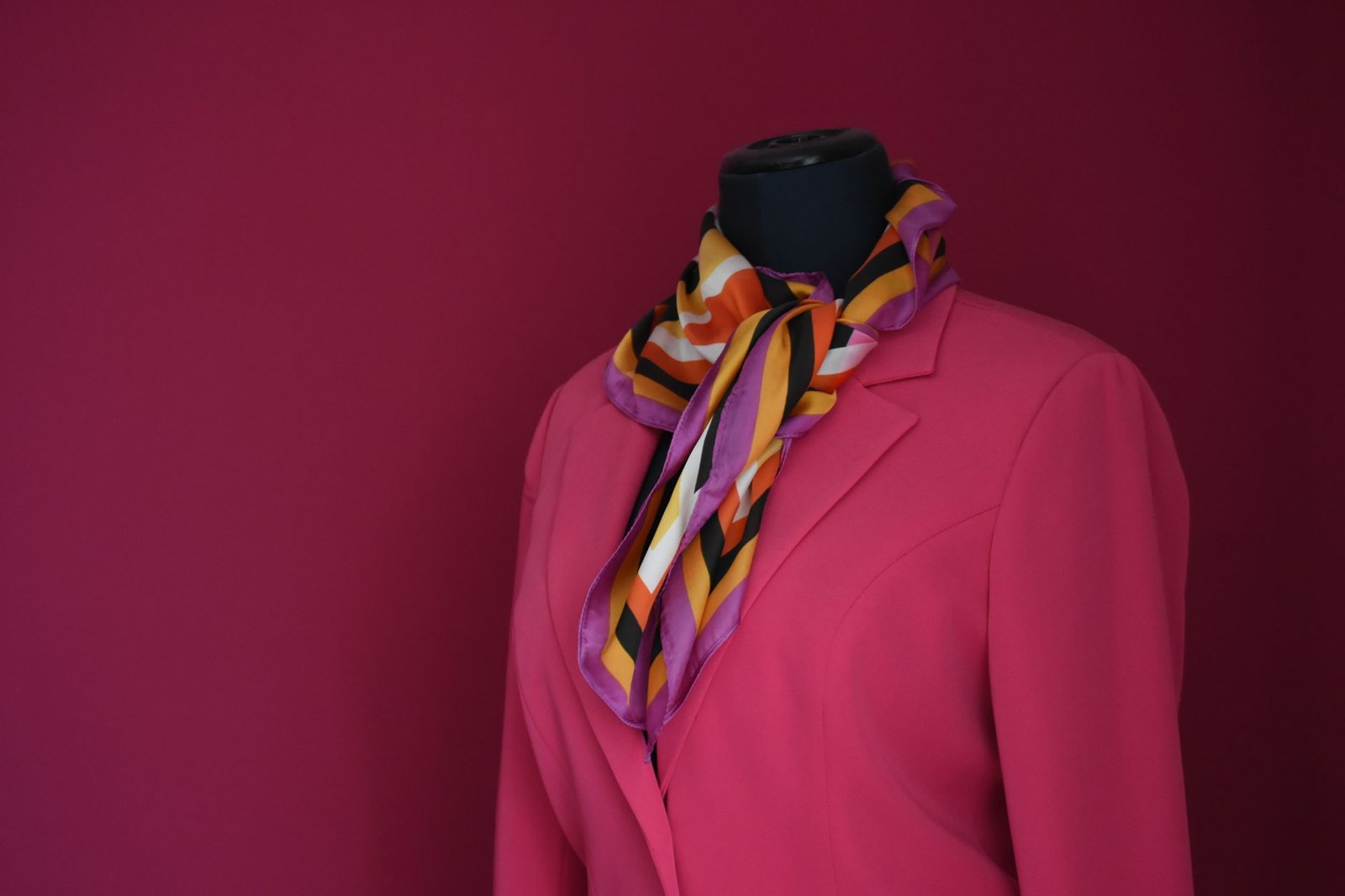 Elegant pink blazer with a colorful scarf displayed on a mannequin against a pink background.