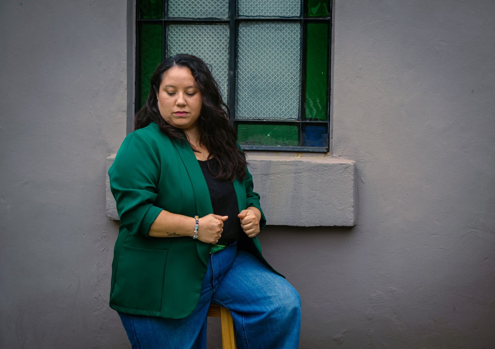 Portrait of a woman sitting on a stool by a stained glass window wearing a green blazer and jeans.