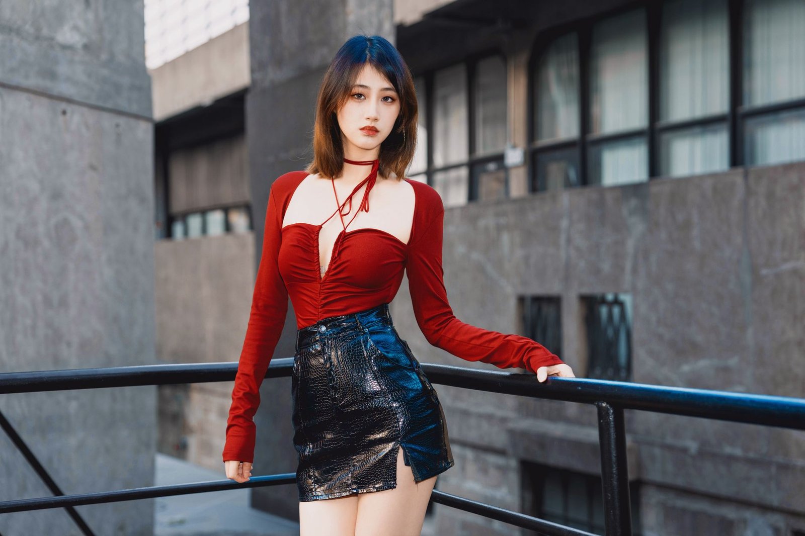 Stylish woman in red top and black leather skirt posing outdoors with modern architecture.