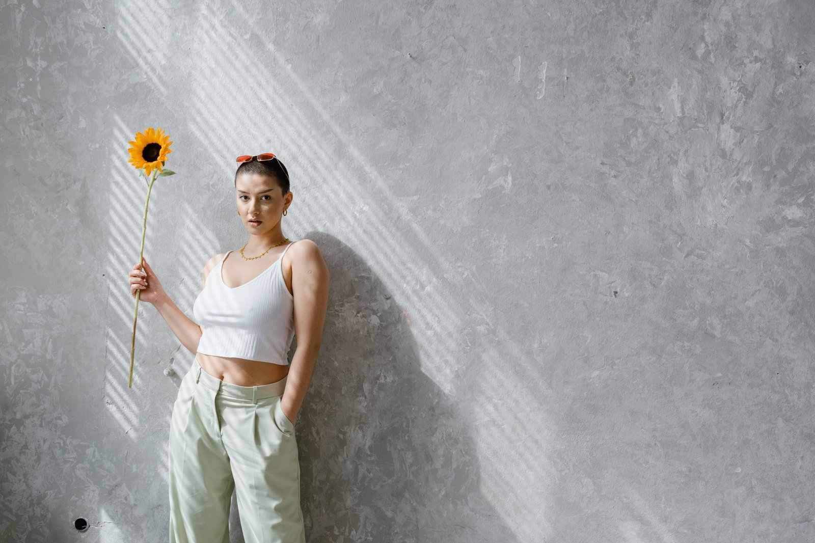 A confident woman in a stylish outfit poses with a sunflower against a textured wall indoors.