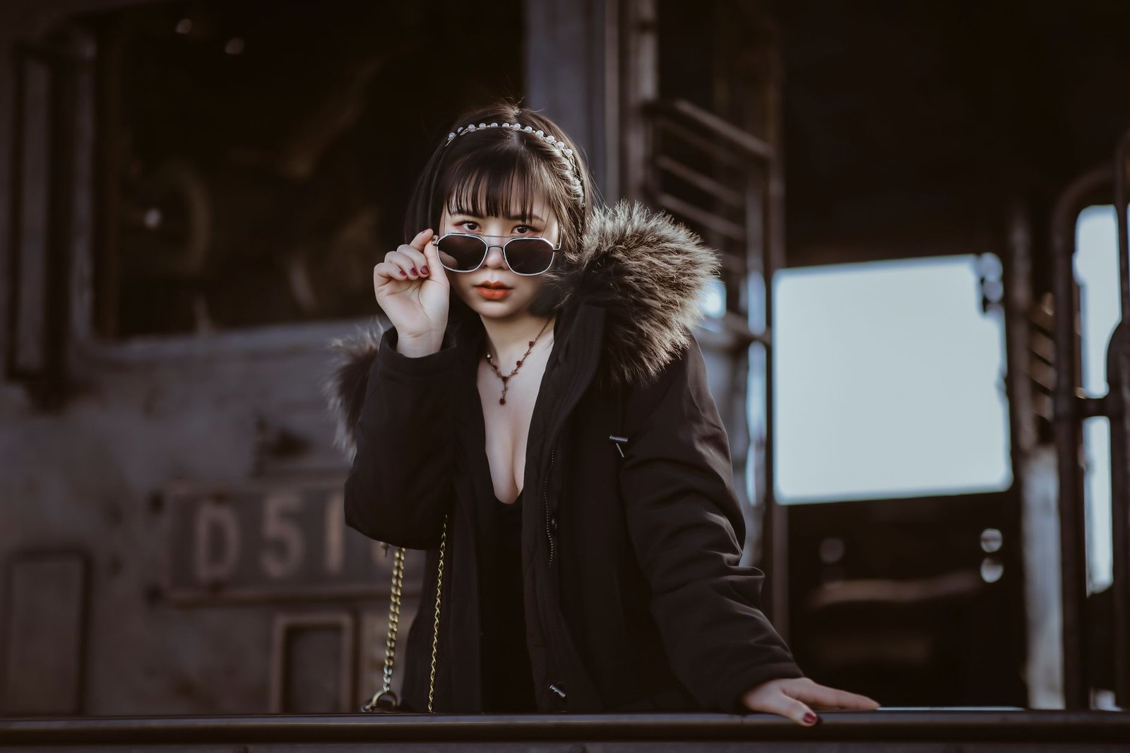 Fashionably dressed woman with sunglasses posing confidently outdoors by an industrial backdrop.