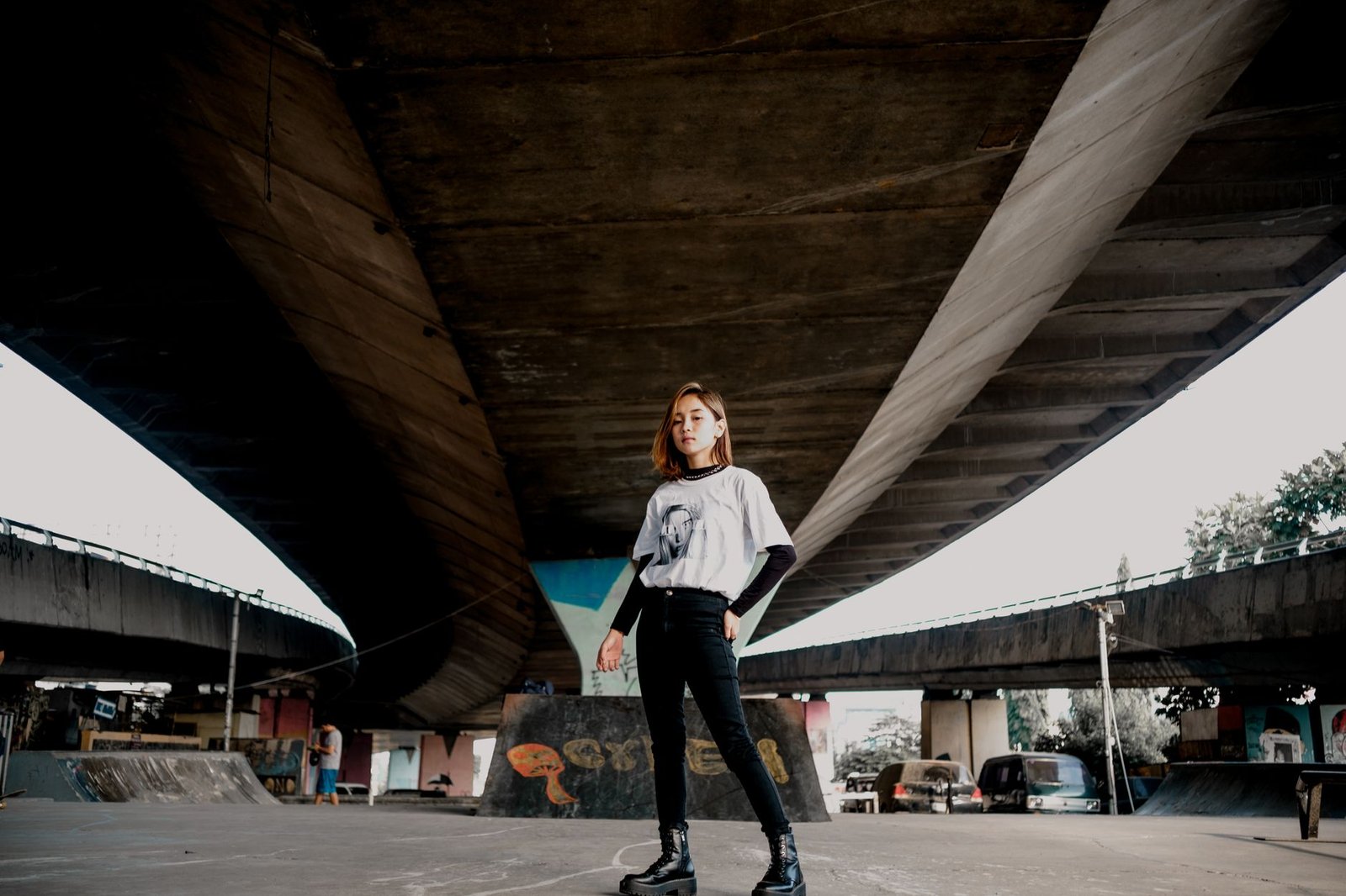 Fashionable young woman stands confidently under an urban bridge.