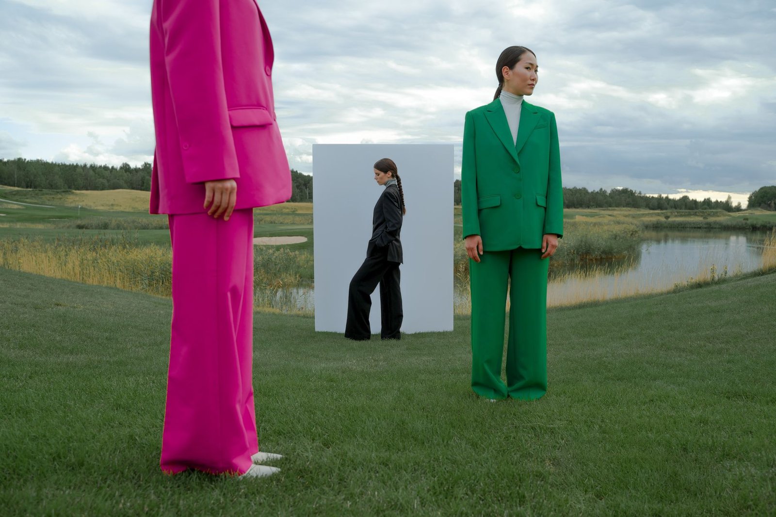 Models in colorful suits pose creatively on a grassy field beside a lake under a dramatic sky.