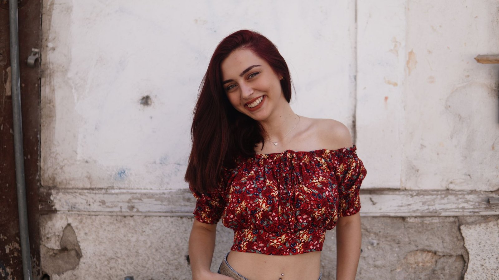 Cheerful female teenager in trendy clothes with naked belly looking at camera near rough wall in town
