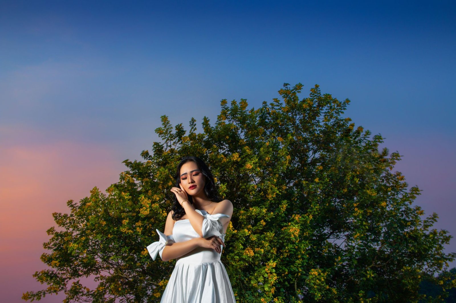 Elegant Asian woman in white dress poses under tree with vibrant twilight sky.