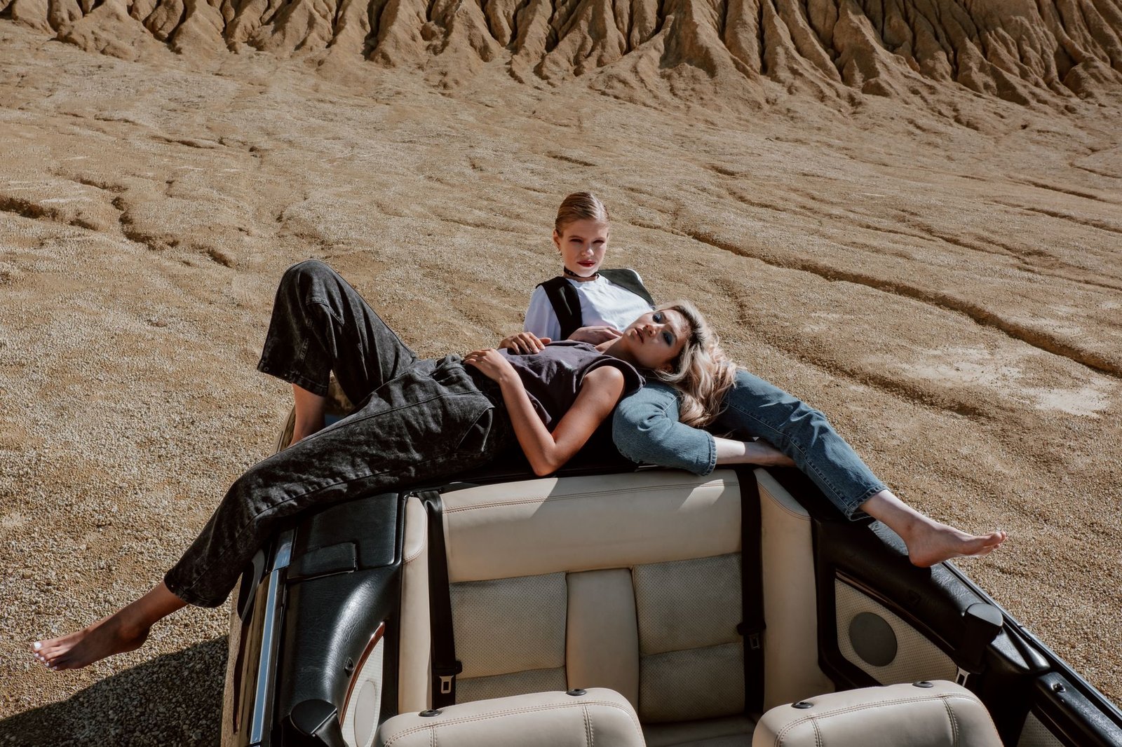 Two women pose stylishly on a convertible in a desert landscape, showcasing modern fashion.