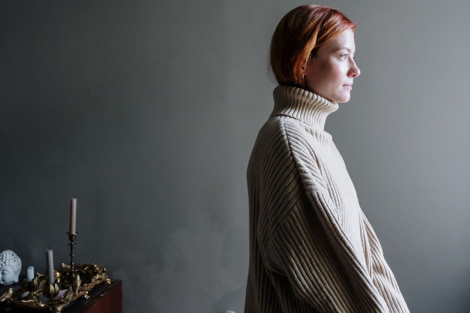 A woman in a ribbed turtleneck sweater stands in a minimalist room with soft lighting.