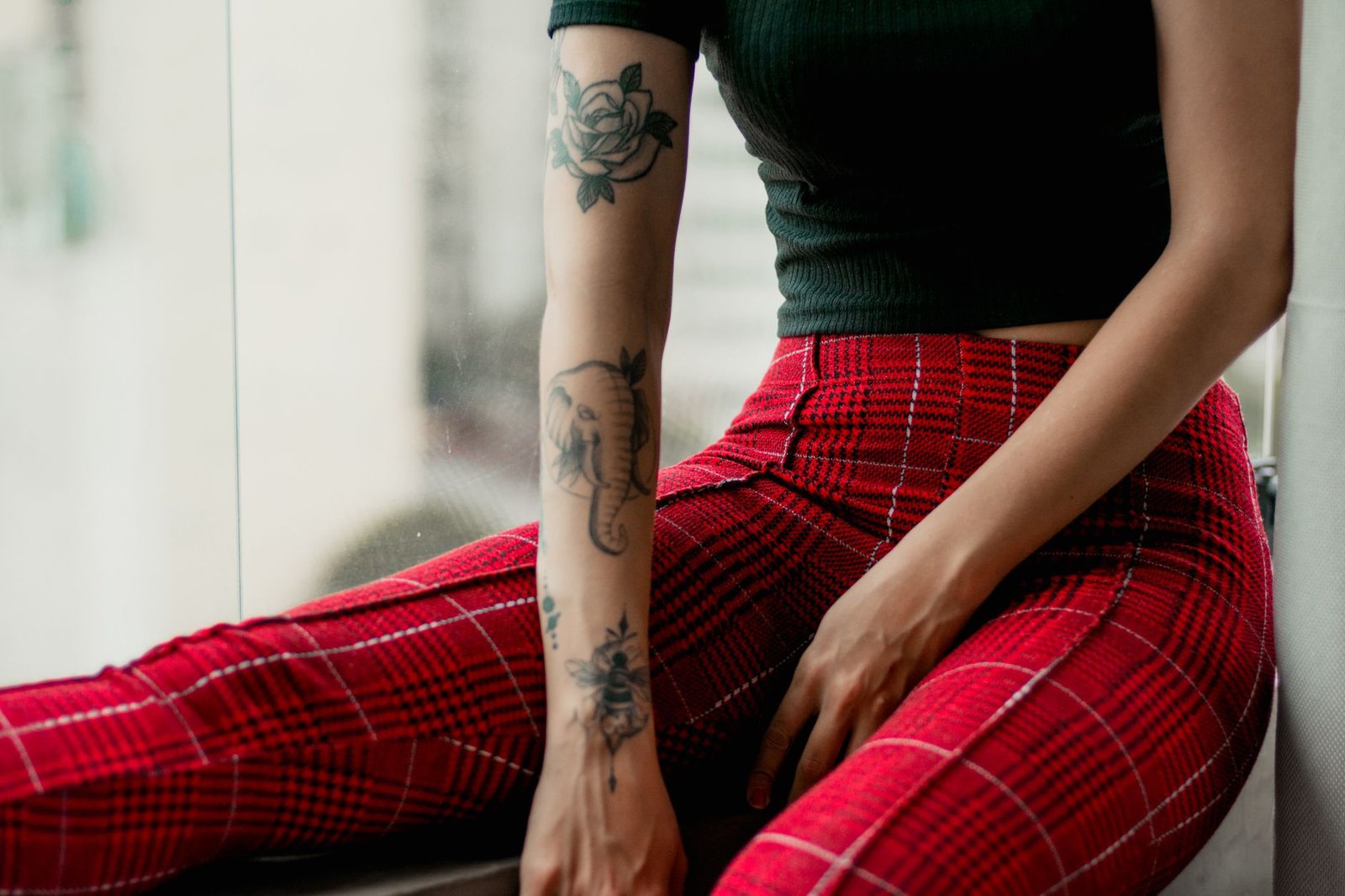 Stylish woman with tattoos poses by a window in red plaid pants, showcasing modern fashion.