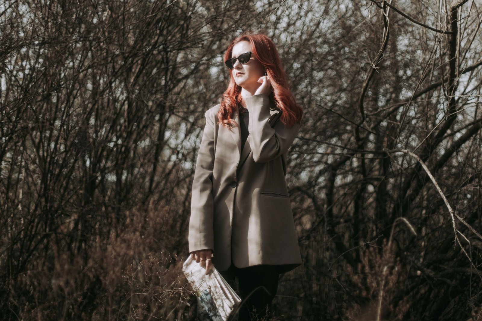 Redhead woman in gray blazer posing with a newspaper in an autumn forest setting.