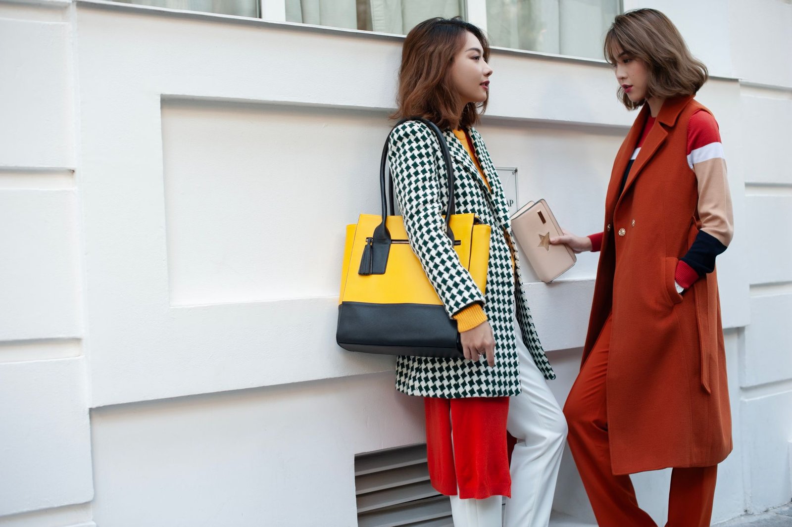 Two fashionable women in bright coats and handbags standing against a white wall outdoors.