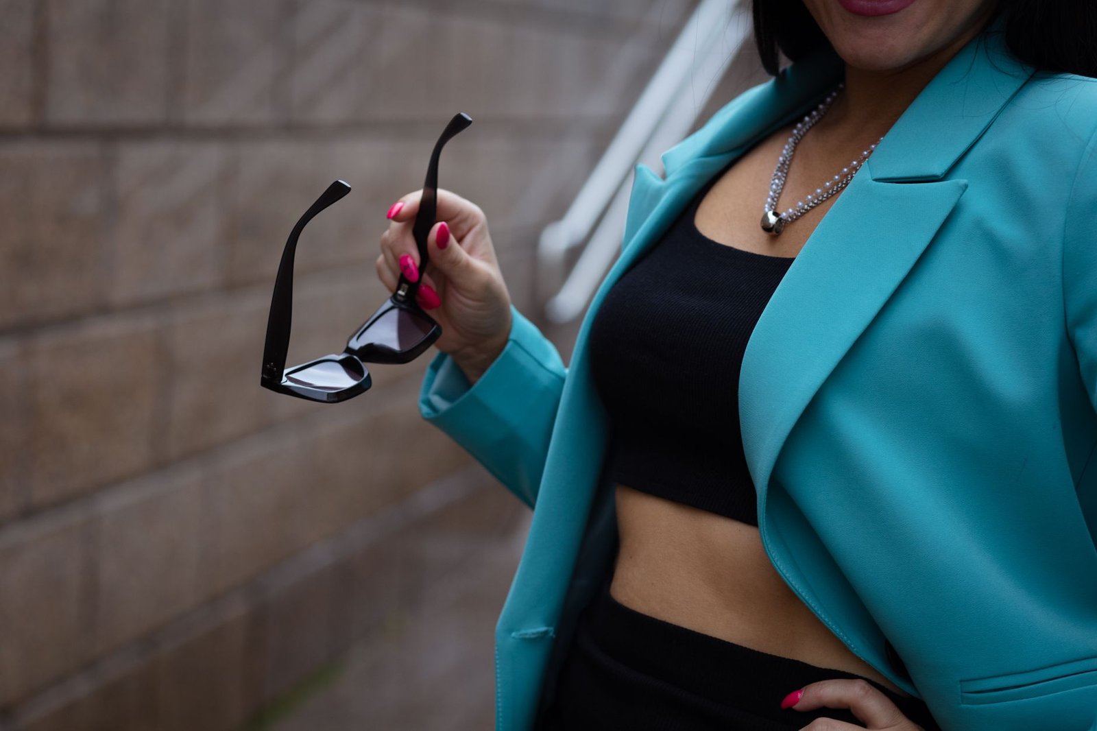 Chic woman in turquoise blazer and crop top holding sunglasses, showcasing urban fashion style.