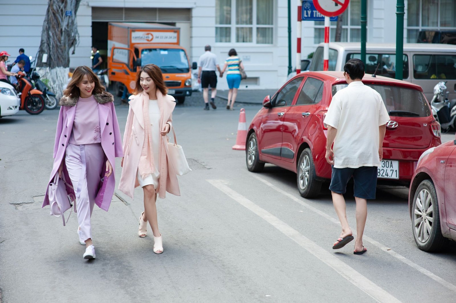 Two stylish women in pastel coats walking on a city street, surrounded by urban life.