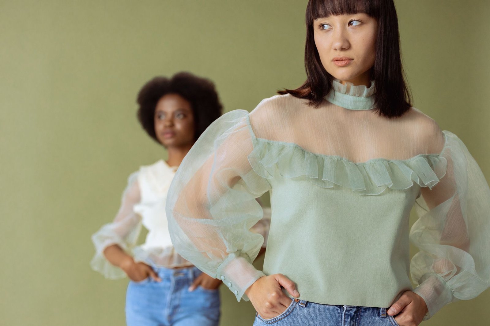 Two women in sheer blouses pose in a stylish studio shot with a subtle green background.