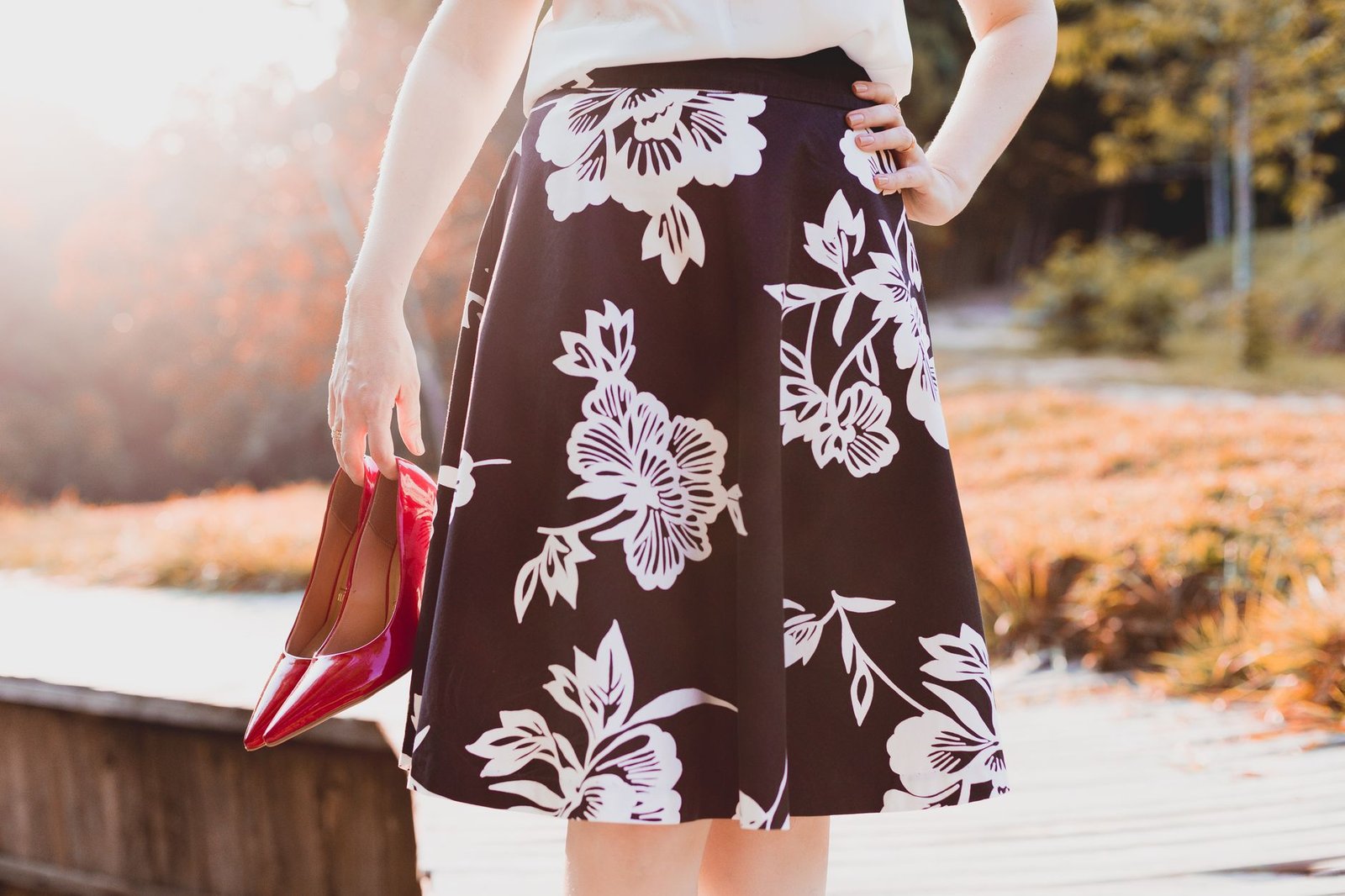 Woman holding red heels, wearing a floral skirt, enjoying a sunny day outdoors.