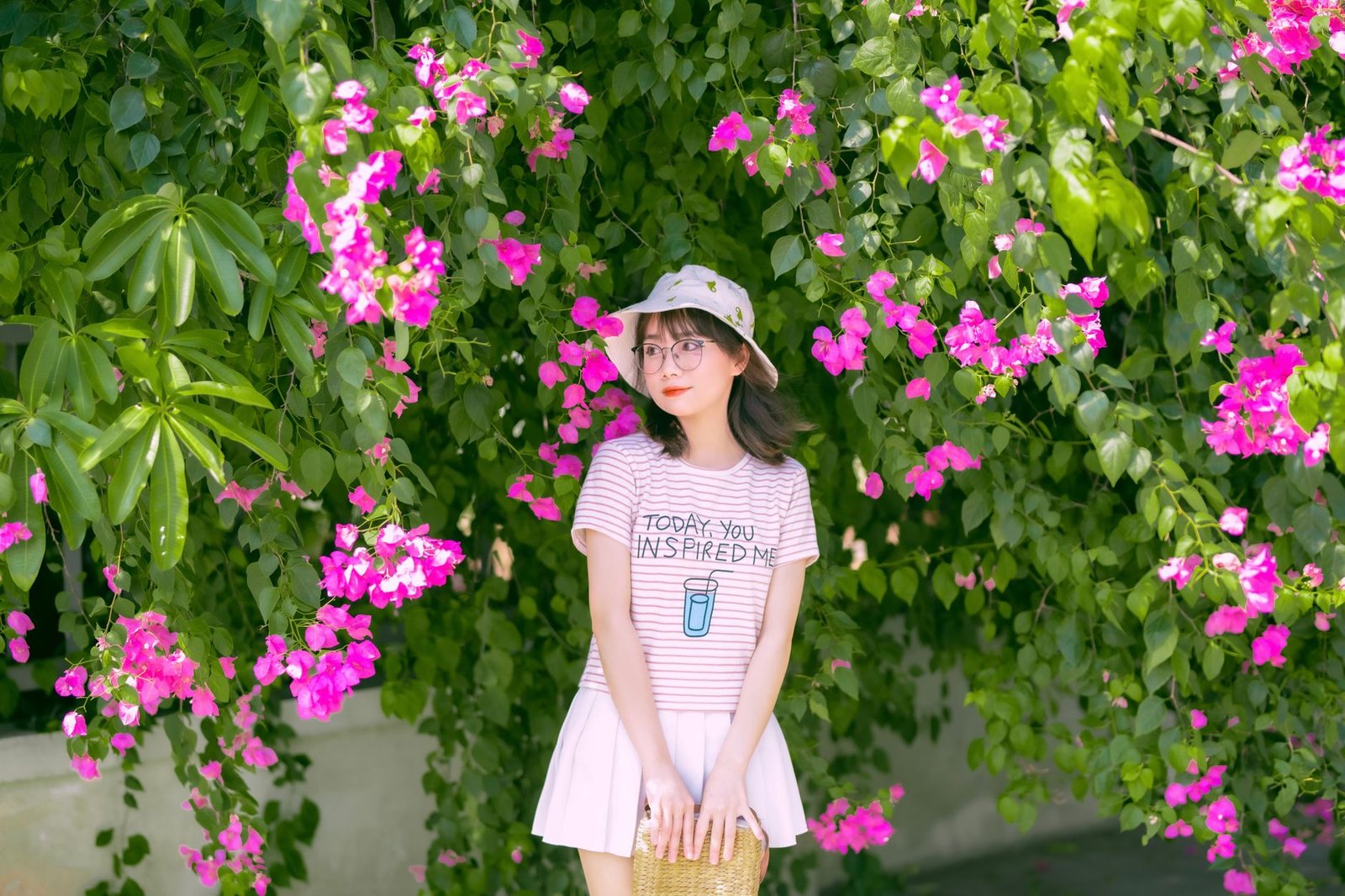 Asian woman standing by colorful bougainvillea, wearing casual fashion with a hat and glasses.