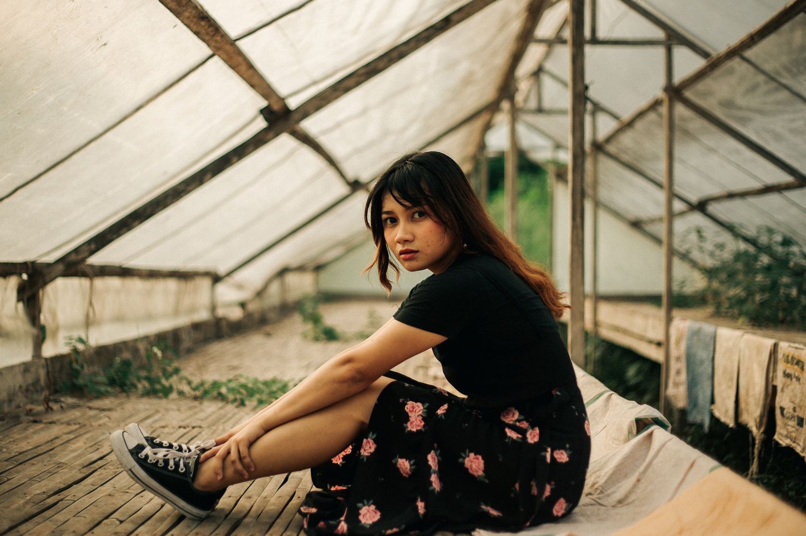 Portrait of a young woman seated in a rustic greenhouse, conveying introspection.
