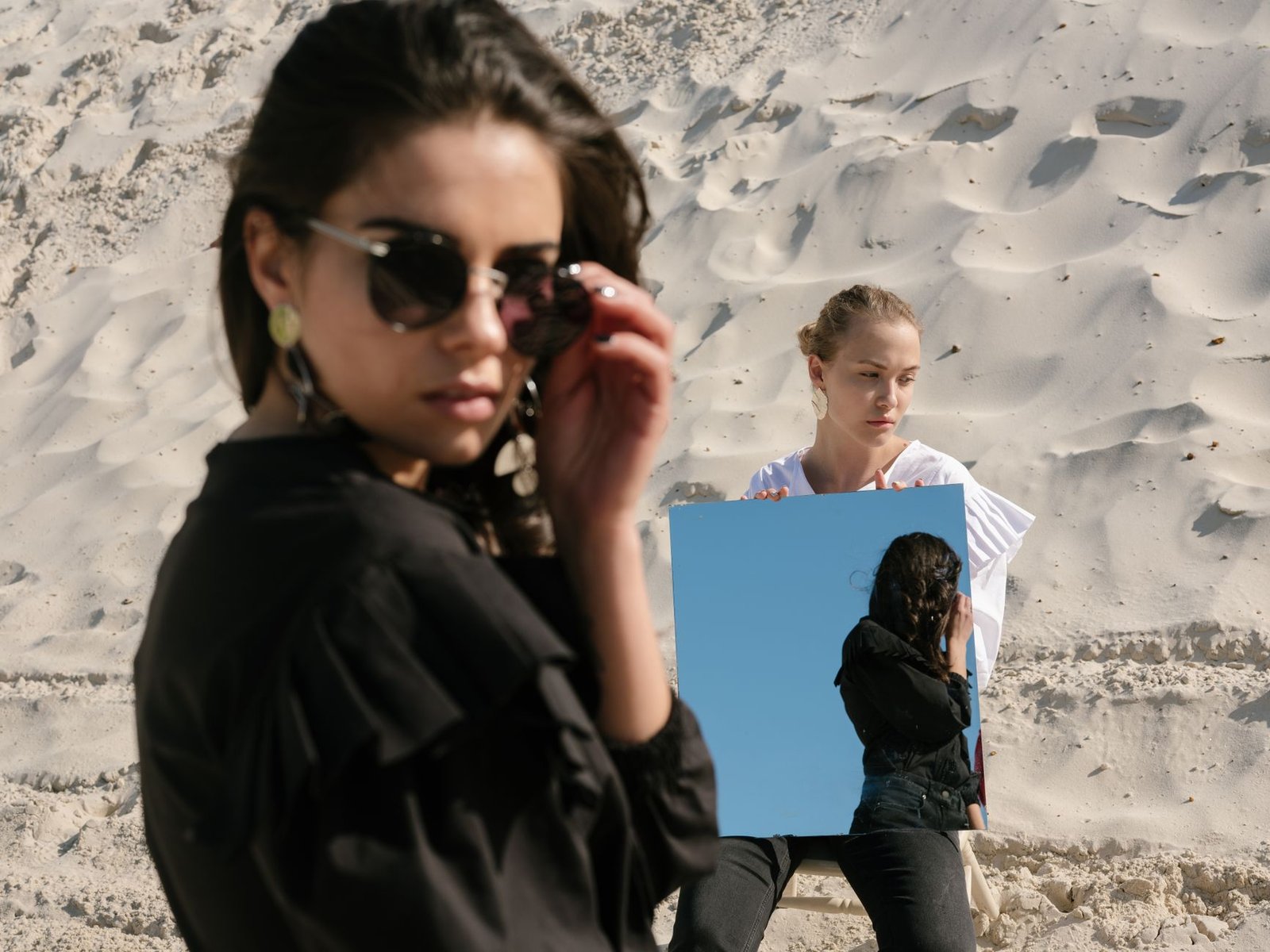 Sad young blond woman sitting on chair with square mirror in hands while attractive brunette female in trendy black outfit and sunglasses reflecting in mirror and looking over shoulder