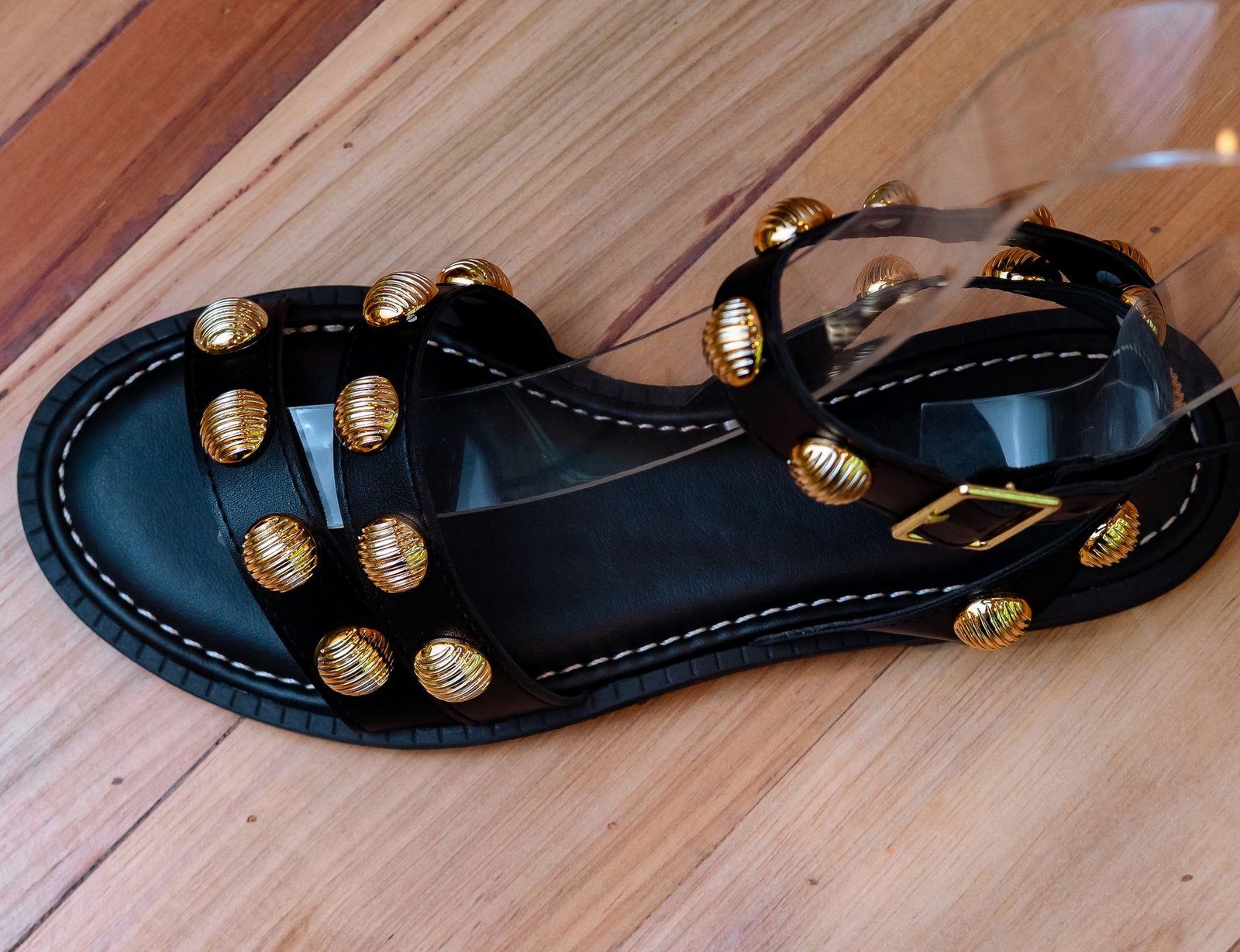 Fashionable black sandal adorned with gold embellishments placed on a wooden floor for a chic look.