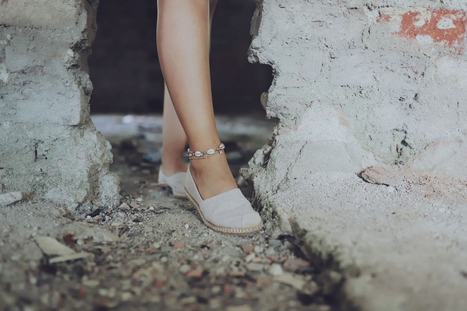Close-up of a woman's leg wearing an ankle bracelet and espadrilles on a rocky, sandy surface.