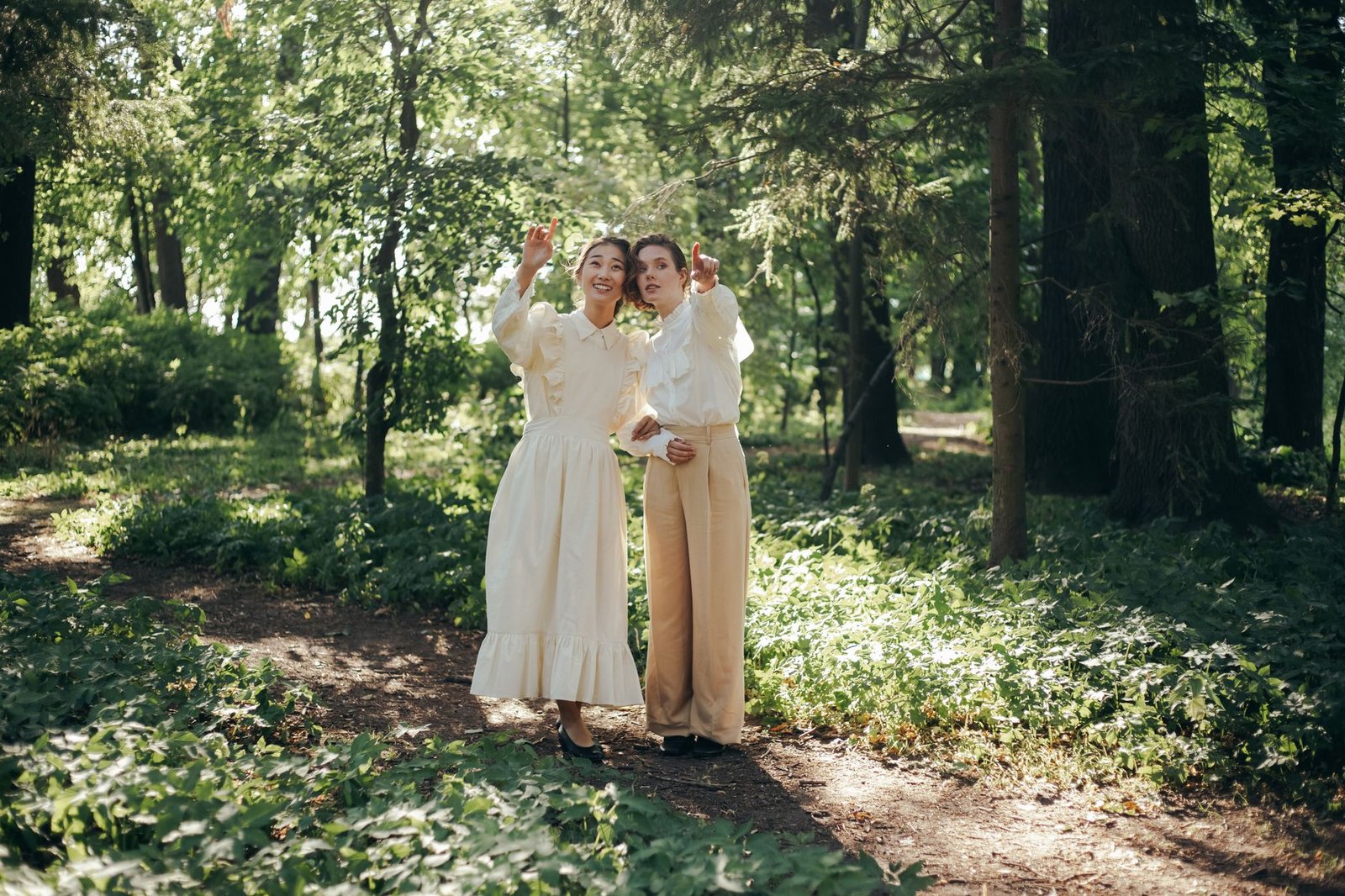 Two women in vintage attire pointing and exploring a lush green forest path.