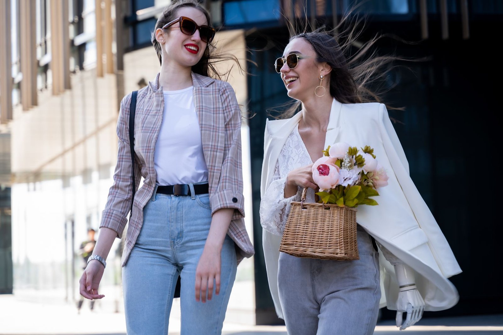 Two stylish women walking outdoors, smiling and enjoying a sunny day with a bouquet of flowers.