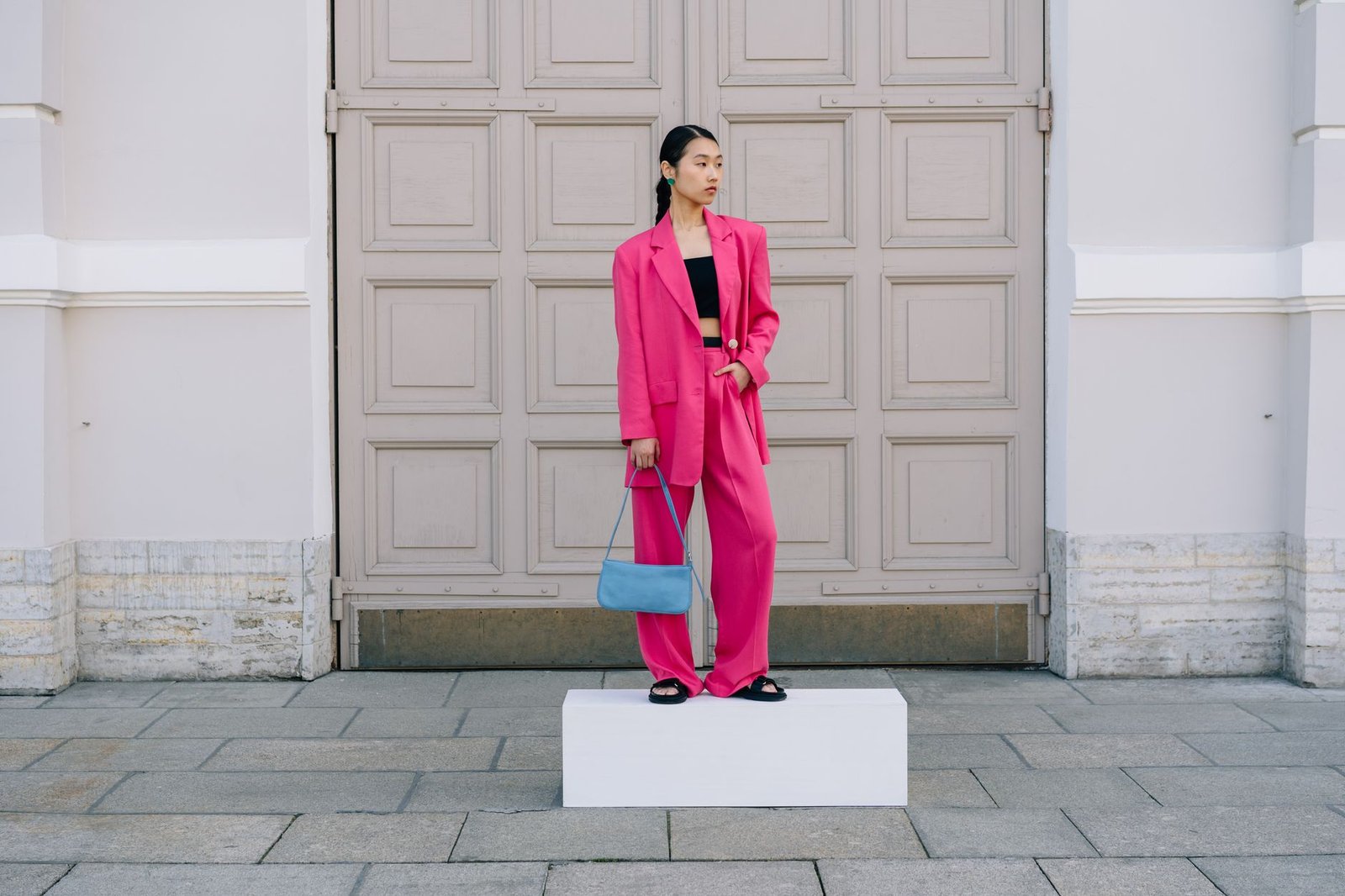 Asian woman in a vibrant pink suit holding a blue handbag, posing confidently outdoors.
