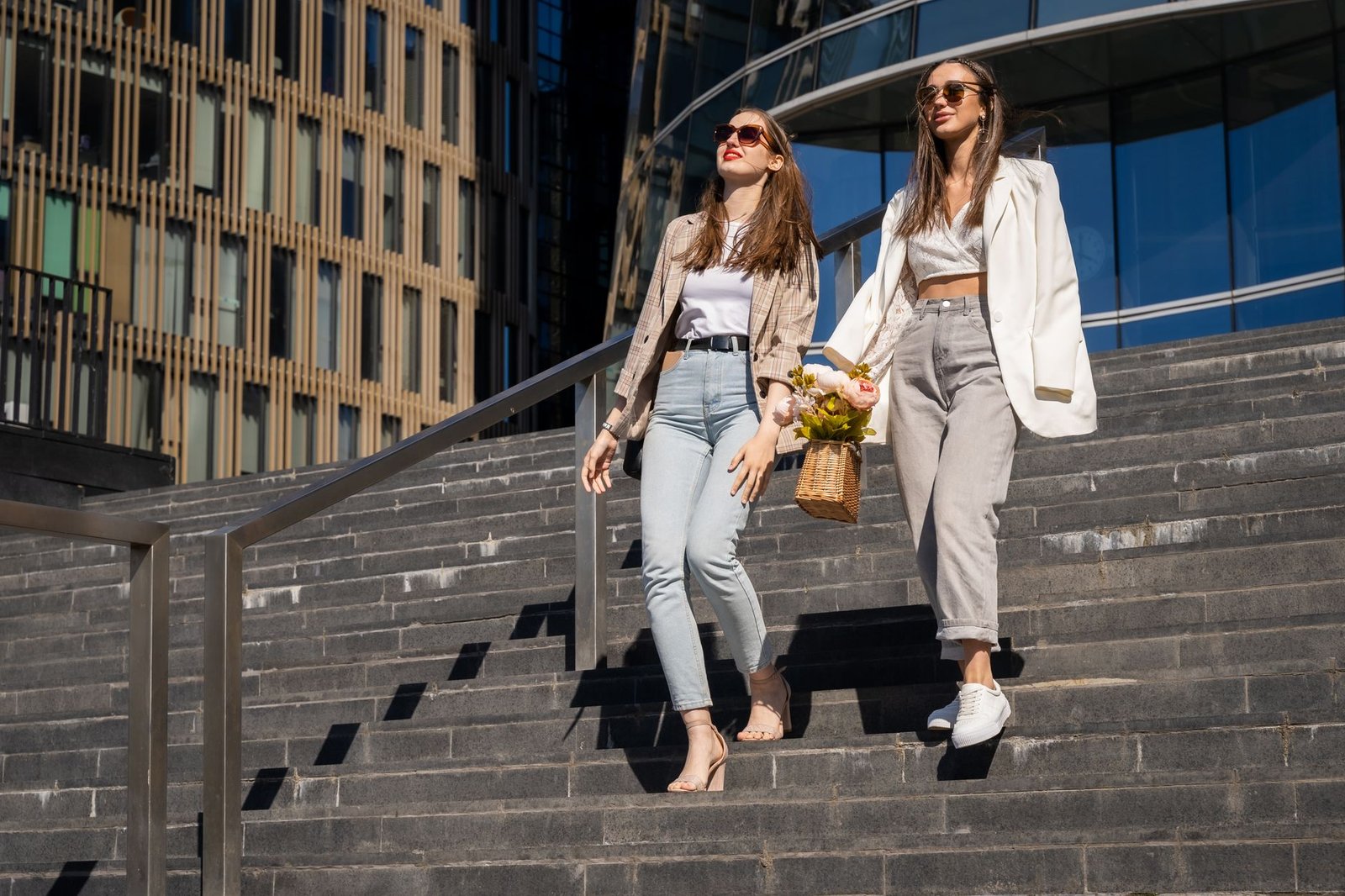 Two fashionable women with sunglasses and a bouquet walk down city steps in the sunshine.
