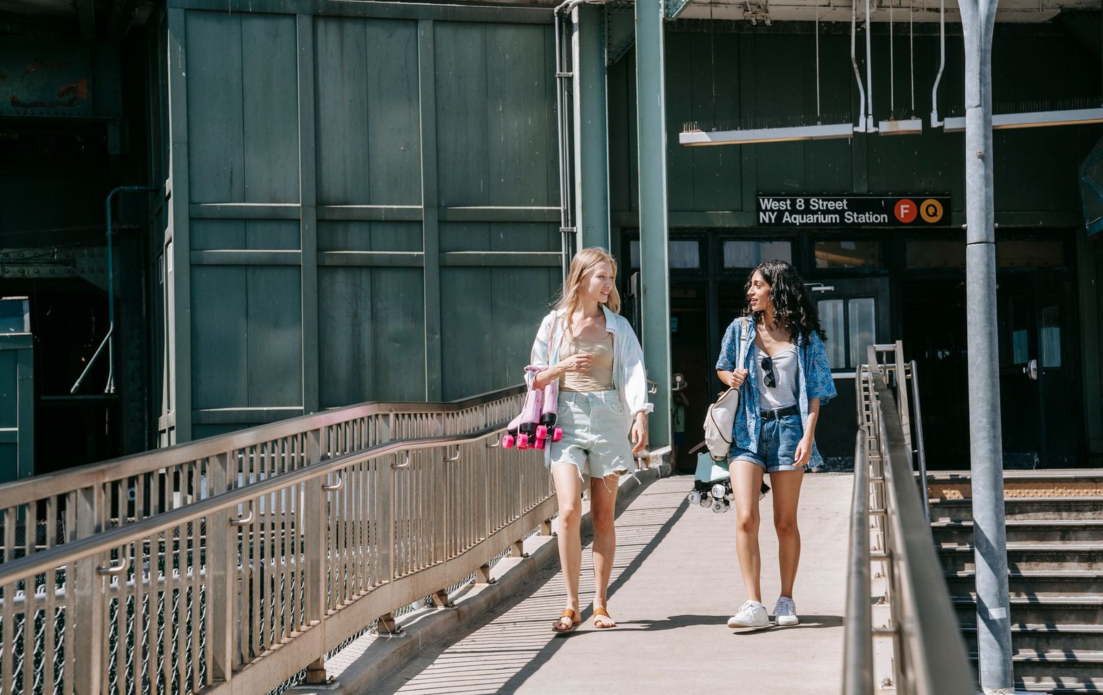 Two young women casually walking on a footbridge outside NY Aquarium Station on a sunny day.