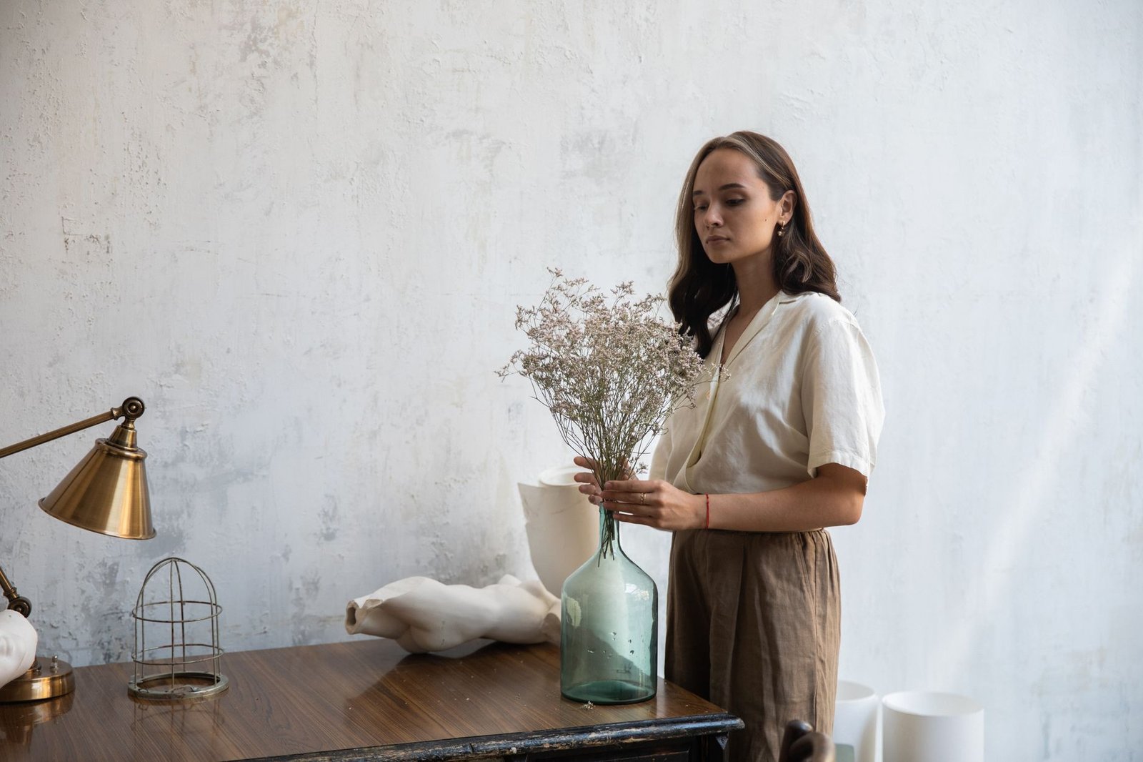 A woman with wavy brown hair arranges flowers in a vase, set in a minimalist, elegant interior with a desk lamp.